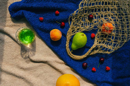 Creative composition made of fruits, glass of lemonede or cocktail and mesh bag on beach towel. Summer refreshment and freshness background. Vegan and vegetarian food concept. Zero west theme. top view. flat layの写真素材