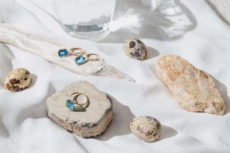 Creative composition made of elegant jewelery set of golden ring and earrings with blue topaz on white background with stones, wood,quail eggs and a glass of water. product still life concept. selective focusの写真素材
