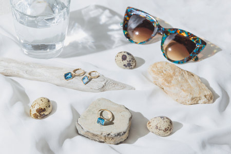Creative composition made of jewelery set of golden ring and earrings with blue topaz on white background with sunglasses, stones, wood,quail eggs and a glass of water. product still life concept. selective focusの写真素材