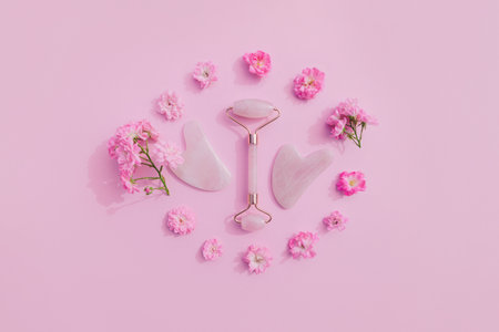 Facial massage kit for home spa. Face roller and gua sha massager made from rose quartz on pink pastel background with rose buds. Natural treatment concept.の写真素材