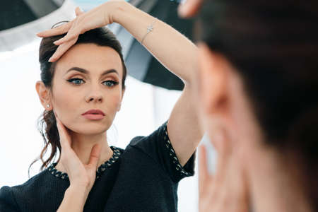 Close up mirror reflection of pretty face of young caucasian brunette woman in little black dress. Woman with blue eyes and nude make-up watches in mirror with studio background.の写真素材