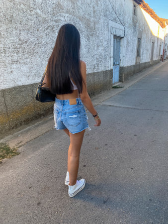Back view of a beautiful young woman with long black hair, wearing jeans shorts and white sneakers, walking along the street.の写真素材