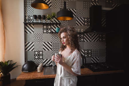 A girl in a light robe is standing in the kitchen holding a cup in her handsの写真素材