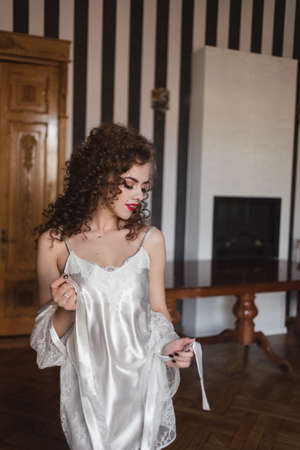 a curly-haired sexy girl standing in a room is dressed in a white robe and is holding a belt from this robeの写真素材