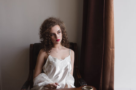 a curly-haired girl sits on a chair near the window, she has a cool figure and she is dressed in a white coat and looks awayの写真素材