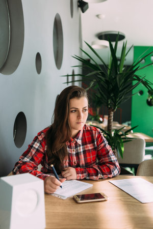 a girl in a red shirt is sitting in a cafe and writing something on paper is lost in thought and looks awayの写真素材
