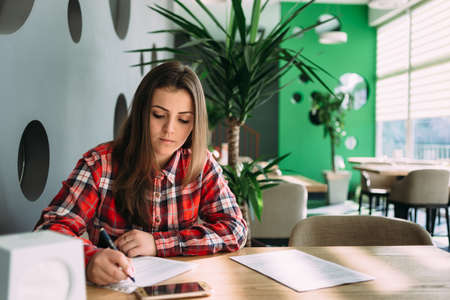 girl student sitting in a bright cafe writing something on paper near her phone lyingの写真素材