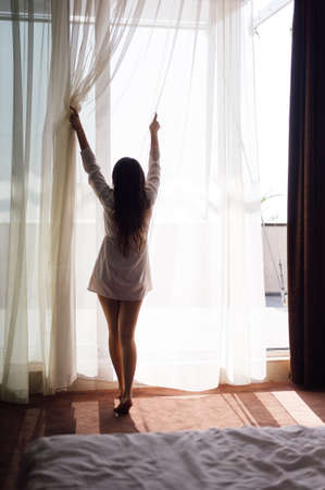 early in the morning a girl stands by the window and looks at him wearing a light robeの写真素材