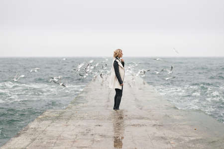 Cute and young woman stands in profile on wet from the waves of the pier. She looks thoughtfully at the sea, touching the collar of her cloak with her left handの写真素材