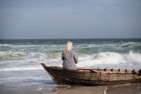 young slender girl sitting alone in a wooden boat by the sea and looking out into the distanceの写真素材