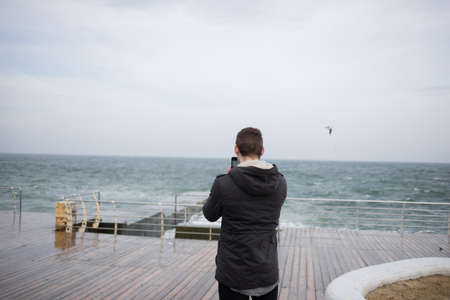 a young guy stands alone on the pier by the sea and takes a photo on the phoneの写真素材