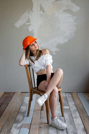 cute girl in the construction orange helmet sits on a chair resting. rest after a hard dayの写真素材