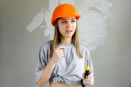 girl on a construction site in an orange helmet with a screwdriver and screw in hand. girl pensively looks away.の写真素材