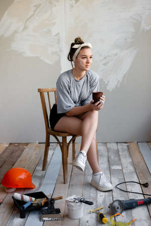 charming girl sitting on a chair drinking coffee resting from repairing a house surrounded by building tools. Tired of repair girl sitting drinking tea.の写真素材