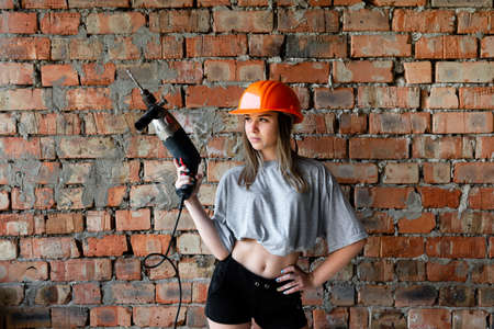 woman in working form in an orange helmet holding a drill on a brick wall background. emancipation. female labor.の写真素材
