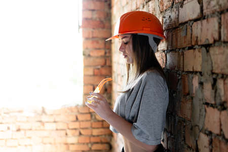 a female construction worker stands by the window leaning on a brick wall in an orange helmet holding protective glasses. the girl has a lunch break at work.の写真素材