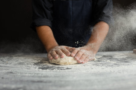the chef kneads raw dough, dust from the flour flies in different directions, male hands on a black...