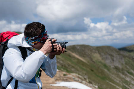 Close up of an adult man taking a photo with his camera at the peak of a mountain with another mountainの写真素材