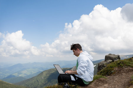 A man works on a laptop sitting on top of a mountain.