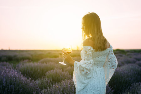 young tender romantic girl stands in a lavender field, in a light dress, hat and holds a glass of French wineの写真素材