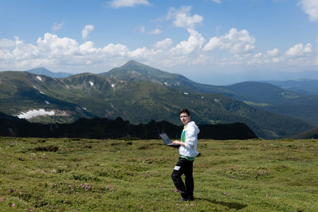 A man with a computer stands in a clearing, mountains in the background