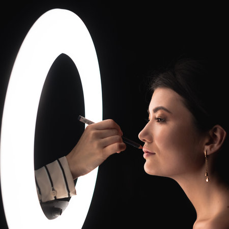 The make-up artist's hand holds a lip pencil and apply straps using a ring lamp for make-up,...