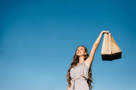 Young cheerful lady holds shopping bag in hands against blue sky.の写真素材