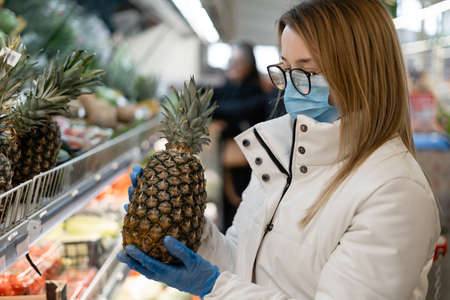 young woman wearing protective equipment buys citruses in a store. pineapples on the shelves of the supermarket. Concept of protecting the population during the COVID-19 pandemic.の写真素材