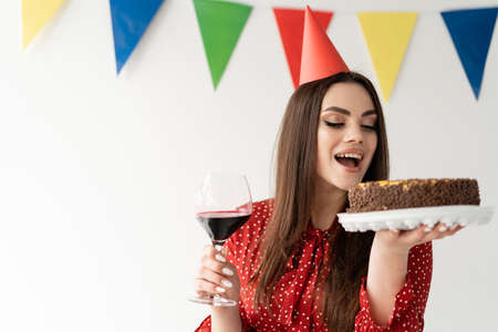 young woman celebrates a birthday wearing a beautiful red dress. Holds a glass of wine and a delicious cake. happy woman. Place for textの写真素材