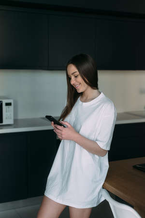 a young woman in a white long T-shirt is standing in the kitchen in her hands holding a mobile phone. Order breakfast online. Online shoppingの写真素材