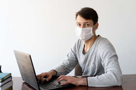 A young man with a medical face shield works on his laptop at home.のeditorial素材