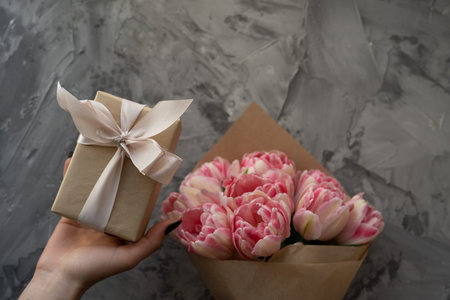 Female hands hold a box with a gift near a bouquet of pink tulips, top view. Place for text. Gift...