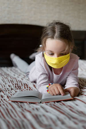 Self-study of school material during quarantine. A little girl lies at home on the couch and reads a book. The concept of distance education.の写真素材