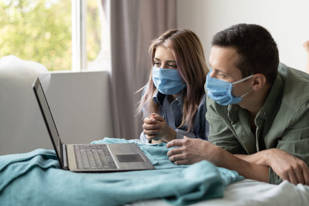 young couple man and woman dressed in medical protective masks lie at home on the bed and watching...