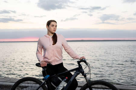 Young beautiful woman, an athlete with a bicycle, from early morning looks at the dawn over the sea and rests from a bike ride.の写真素材