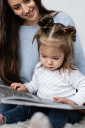 A little girl two years old is sitting with her mother on her lap. Stay at home. Mom with daughter reads a big paper book.の写真素材