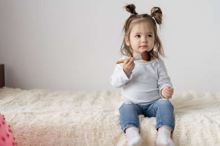 a little girl of European appearance sits at home on the bed. The child is 2 years old. Powder brush in the hands. Play gamesの写真素材