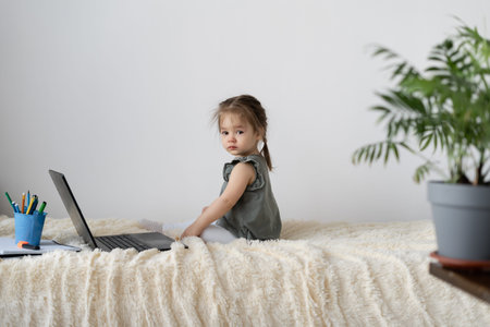 A little girl of European appearance sits at home on the bed. Plays online in a laptop.