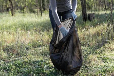 in the forest, a volunteer collects garbage in a black bag. Female hand in a rubber glove. To clean...