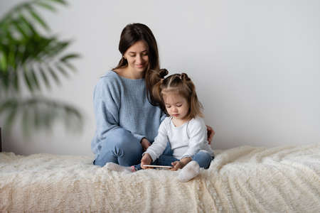 A little girl two years old is sitting on the bed with her mother. Play games on a mobile phone. Smartphone in the hands of a child. White wall place for text.の写真素材