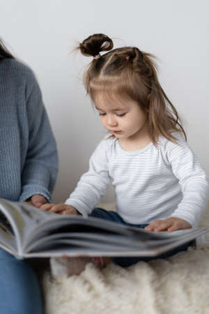 A young mother sits on the bed with her two-year-old daughter and read a paper book together. A girl is watching drawings.の写真素材