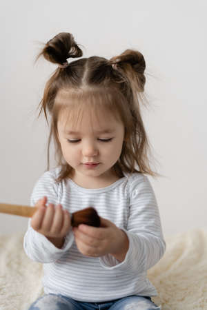 Portrait of a little girl three years old. Sits on the sofa holding a makeup brush in his hand. The child is playing a game. Imagines himself a stylist. Dreaming about a professionの写真素材