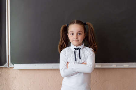 A young girl is standing at the blackboard. School education. Portrait of a blond child at school. Parenting and education of children. Hands on the chest of the girl.の写真素材