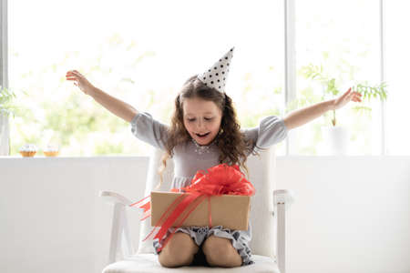 little girl sitting at home on the armchair near the window. Birthday at the child. Holiday and joyful gift. Emotions of delight and joy at the birthday girlの写真素材