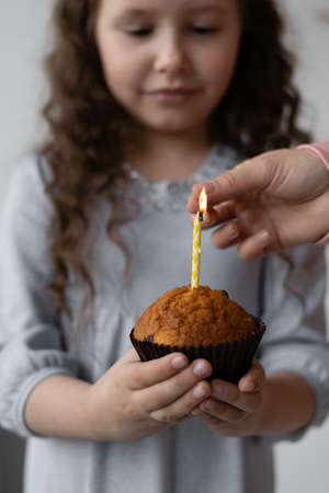 Cute girl holding a cupcake in her hands with one candle, part of her body, happy holiday, birthday, fun and joy concept. Mom sets fire to a candleの写真素材
