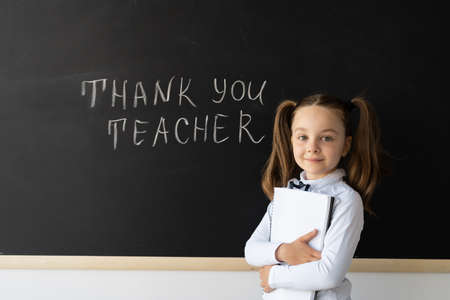 Little girl schoolgirl stands at the blackboard. She has in her hands a large school notebook. She wrote on the blackboard. Education. Study. School.の写真素材