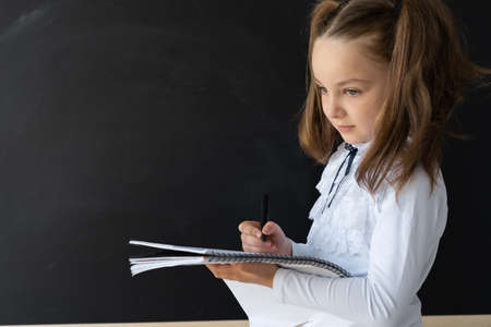 Girl schoolgirl stands in front of the blackboard. She makes notes in a notebook with a pencil. She is wearing a school uniform.の写真素材