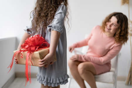 Birthday concept. Happy young woman with wavy hair is sitting in a chair. The girl hides her hands with a gift behind her back and does not show her mom.の写真素材