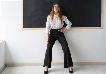 Smart and beautiful teacher in her office. She is standing in front of a black board. Higher education. The lady is wearing a sexy white shirt and black pants. Black modern shoes on his feet. One of her hands on the belt hangs the second along the body.の写真素材