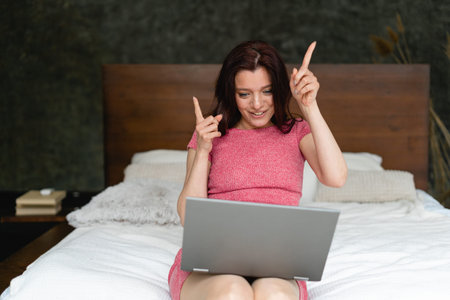 A young woman in pajamas is sitting on the bed in the room and gesturing. On her feet lies a laptop....